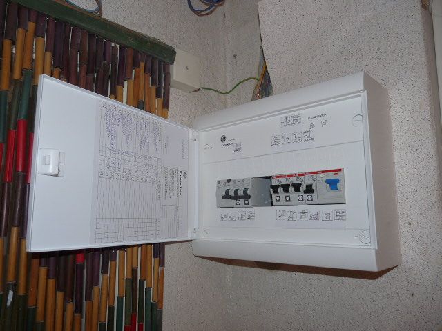 Tableau électrique blanc à couvercle ouvert, fixé au mur près de rideaux colorés.