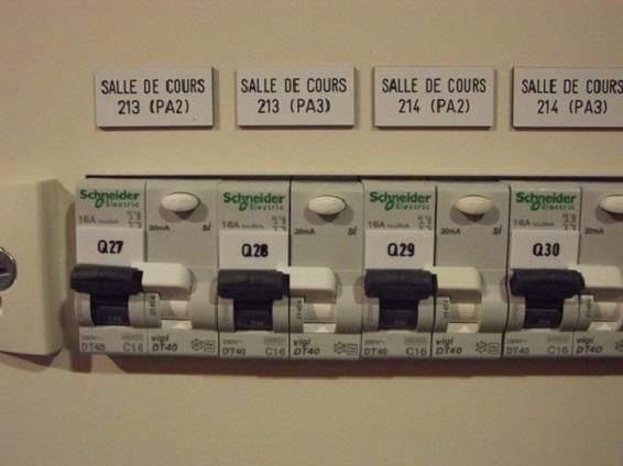 Rangée d'interrupteurs de disjoncteurs électriques étiquetés pour les salles de classe 213–214, avec des étiquettes de salle écrites à la main au-dessus.