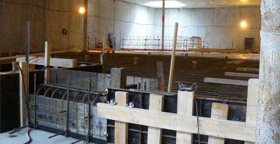 Grand chantier de construction intérieur avec fondations en béton, armatures et coffrages en bois sous une lumière vive
