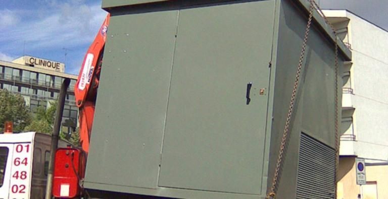 Une grande armoire utilitaire verte est soulevée par une grue rouge à l'extérieur, près d'un bâtiment.