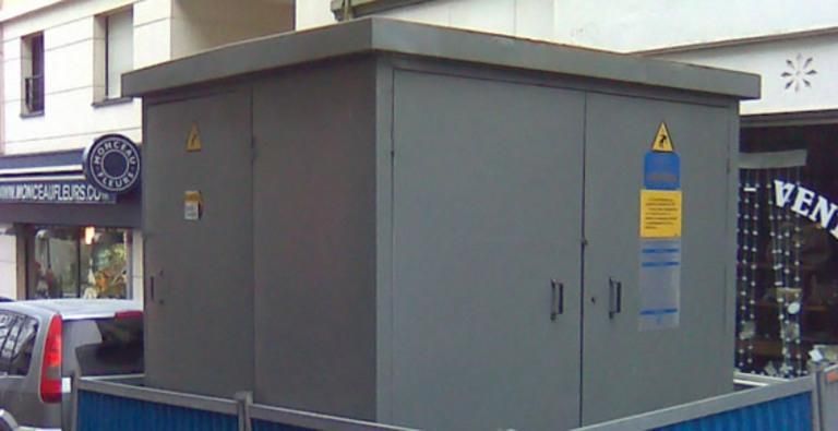 Boîtier électrique gris sur un trottoir, à côté d'une vitrine et de voitures stationnées.