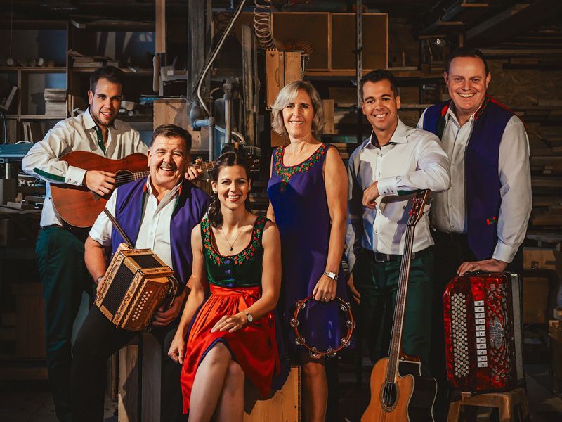 Sechs Musiker in traditioneller Kleidung posieren in einer Werkstatt; einer mit Gitarre, Akkordeon und Tamburin.