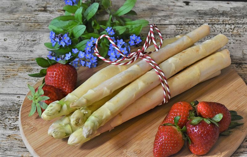 Ein Bündel weißer Spargel, mit einer Schnur zusammengebunden, liegt neben Erdbeeren und blauen Blumen auf einem Holzbrett.