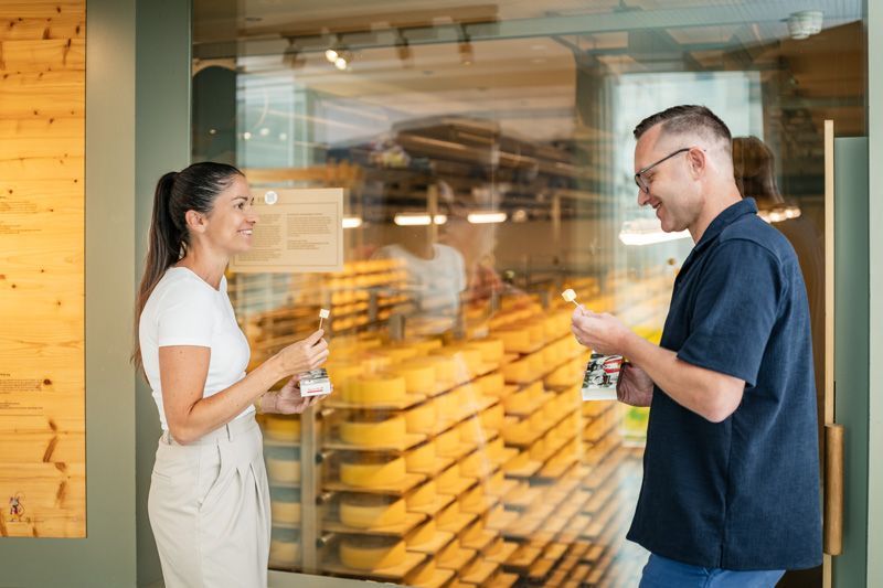Frau und Mann vor einem Käsekeller, halten Käseproben in den Händen und lächeln. Holzvertäfelung und großes Glasfenster.