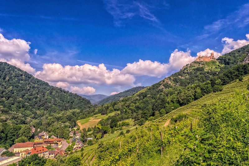Grüne Berge mit einem Tal, einem Dorf und einer Burg auf einem Hügel unter blauem Himmel mit flauschigen Wolken.