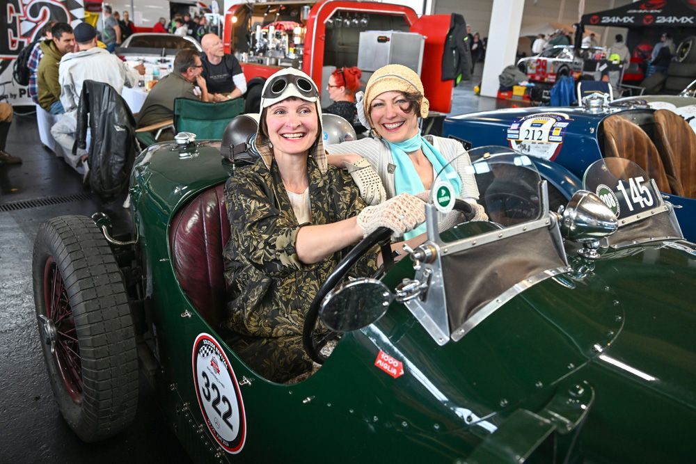 Zwei lächelnde Personen in Oldtimer-Fahrerkleidung sitzen bei einer Indoor-Veranstaltung in einem grünen, klassischen offenen Rennwagen.
