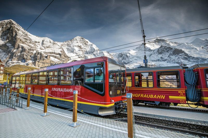 Ein roter Zug der Jungfraubahn steht an einem Bahnhof mit schneebedeckten Bergen im Hintergrund.
