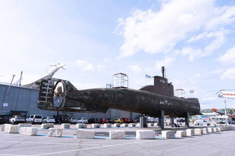 Ein dunkles U-Boot, gestützt von Betonblöcken, steht auf einem Parkplatz unter bewölktem Himmel.