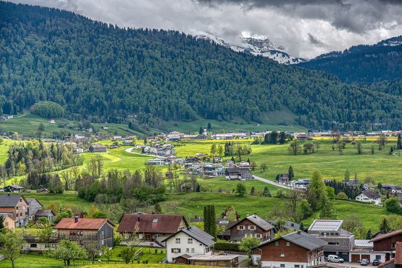 Grünes Tal mit Dorf, umgeben von bewaldeten Bergen unter bewölktem Himmel.