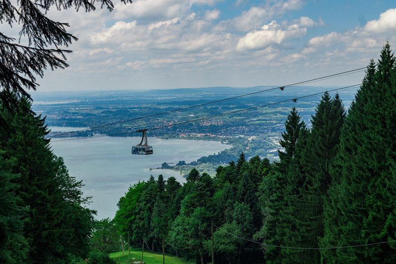 Seilbahn über einen See, Blick von einem bewaldeten Hang aus. Blauer Himmel, grüne Bäume, Stadt in der Ferne.