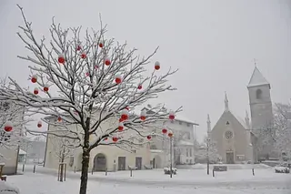 Ein schneebedeckter Baum mit roten Kugeln darauf vor einer Kirche.