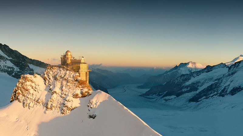Ein schneebedeckter Berggipfel mit einem Gebäude darauf, goldenes Sonnenlicht erhellt die Landschaft.
