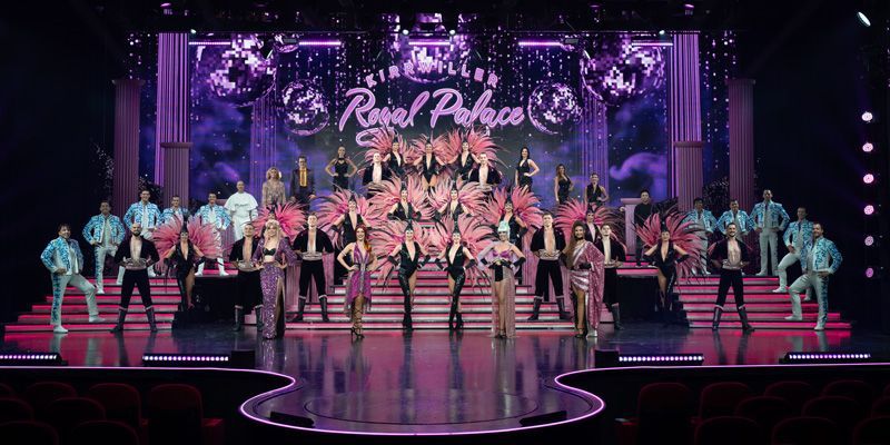 Eine große Gruppe von Künstlern steht mit einem Schild mit der Aufschrift „Königlicher Palast“ auf der Bühne. Die Bühne ist in Rosa und Lila beleuchtet.