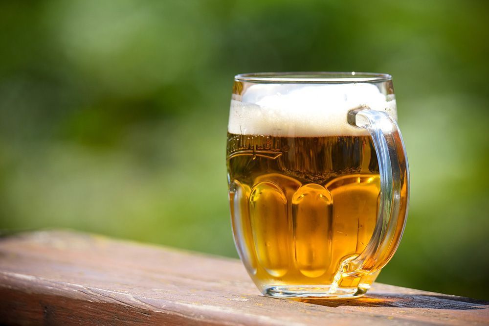Ein mit goldenem Bier gefüllter Glaskrug mit schaumiger Krone steht auf einer Holzoberfläche vor einem verschwommenen grünen Hintergrund.
