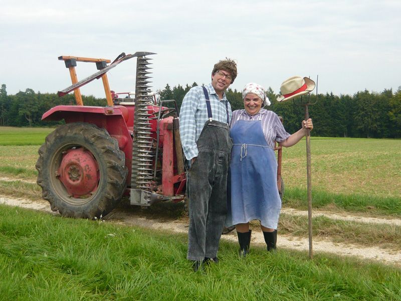 Ein Mann und eine Frau in Bauernkleidung stehen neben einem roten Traktor auf einem Feld. Die Frau hält einen Gehstock.