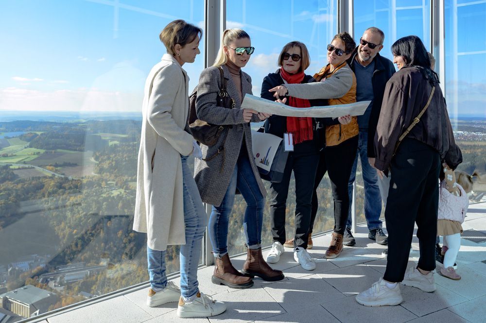 Eine Gruppe von Menschen steht auf einer Aussichtsplattform in einem Hochhaus und betrachtet gemeinsam eine Karte vor dem Hintergrund einer malerischen Landschaft.