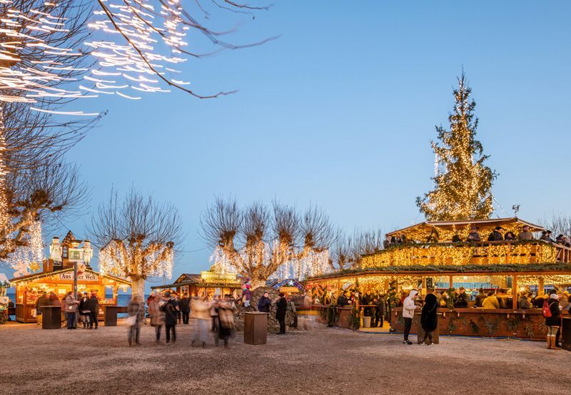 Weihnachtsmarktplatz mit beleuchteten Ständen, Karussell und Weihnachtsbaum in der Abenddämmerung.