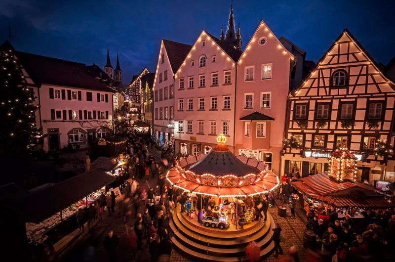 Nachtansicht eines festlich geschmückten europäischen Stadtplatzes mit Karussell, beleuchteten Gebäuden und einer Menschenmenge, die sich zum Weihnachtsmarkt versammelt hat.
