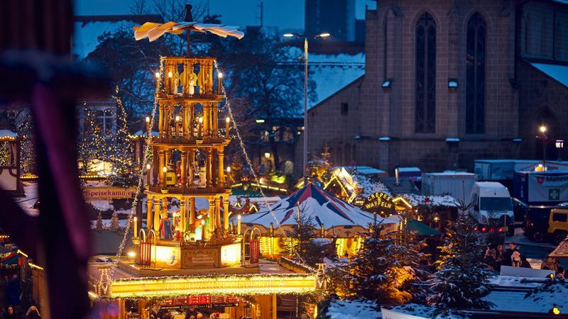 Weihnachtsmarkt in der Abenddämmerung mit beleuchteter Pyramide und Karussell, in der Nähe einer Kirche.