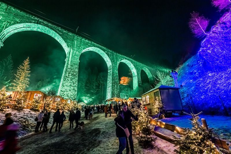 Nachtszene mit beleuchteten Steinbögen und darunterliegenden Weihnachtsmarktständen. Menschen gehen im Schnee.
