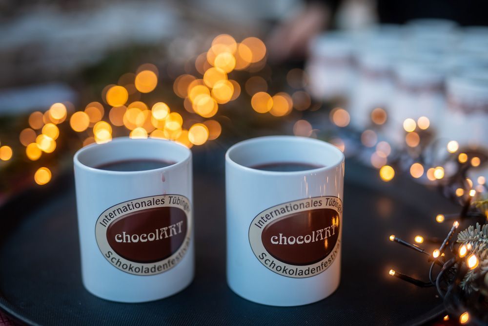 Zwei Becher mit der Aufschrift „chocolART“ auf einem Tablett vor dem Hintergrund festlicher, unscharfer goldener Lichterketten.