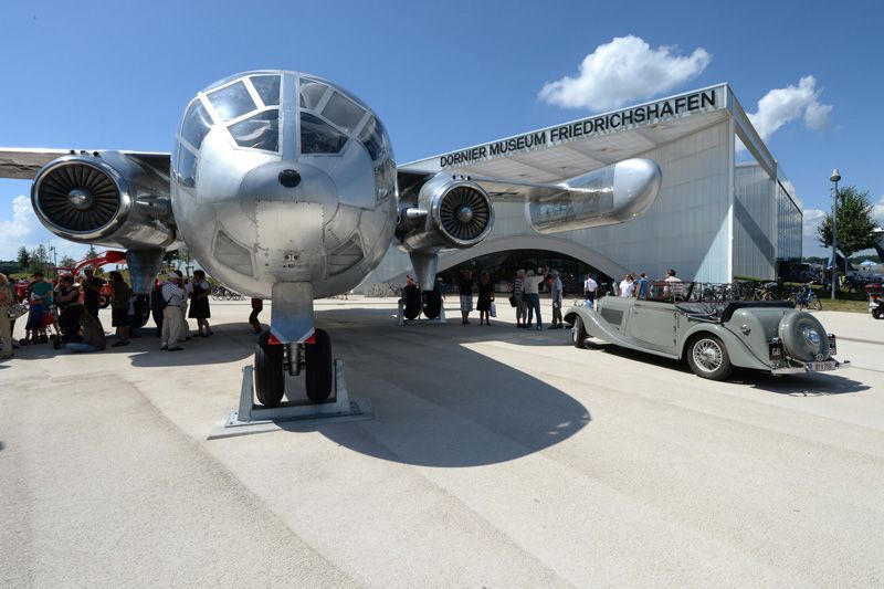 Vor einem Museumsgebäude ist ein großes silbernes Flugzeug ausgestellt, davor parkt ein Oldtimer.