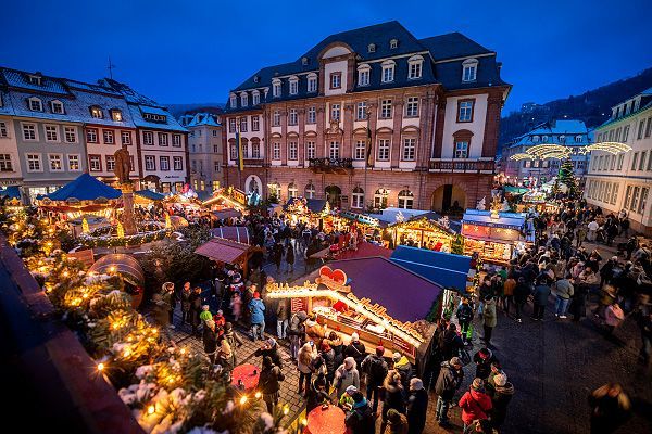 Weihnachtsmarkt in der Abenddämmerung, Heidelberg, Deutschland. Menschen schlendern an den beleuchteten Ständen entlang; im Hintergrund historische Gebäude.