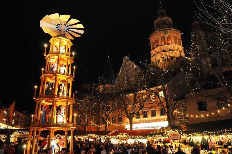 Weihnachtsmarkt bei Nacht, im Hintergrund eine beleuchtete Holzpyramide und eine Kirche. Viele Menschen.