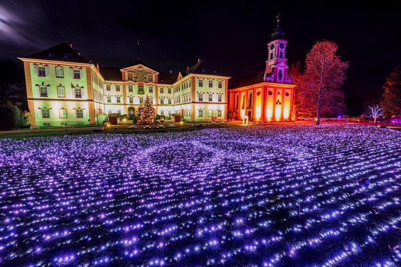Nachts beleuchteter Palast und Kirche, davor ein Feld aus violetten Lichtern.