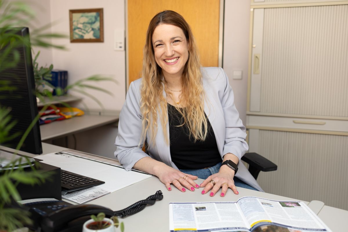 Eine Frau sitzt an einem Schreibtisch vor einem Computer und lächelt.
