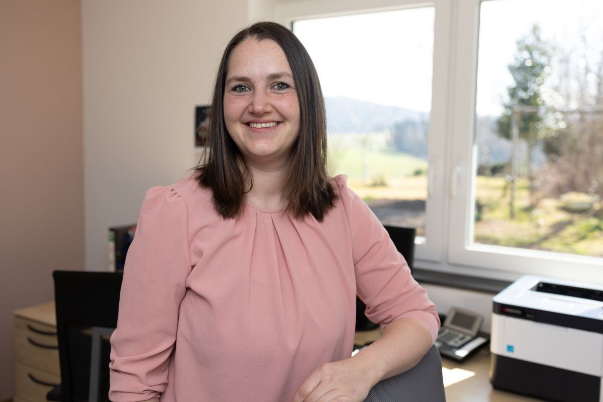 Eine Frau in einem rosa Hemd sitzt an einem Schreibtisch vor einem Fenster.