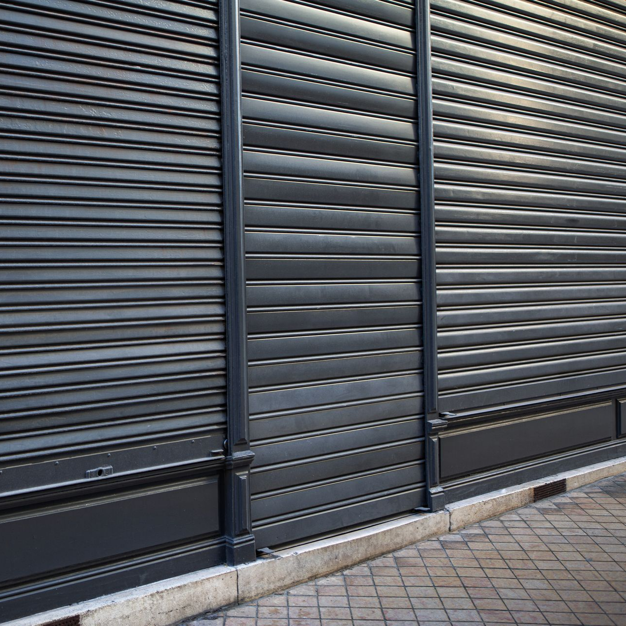 Volets de sécurité en métal gris devanture de magasin, fermés, sur un trottoir.