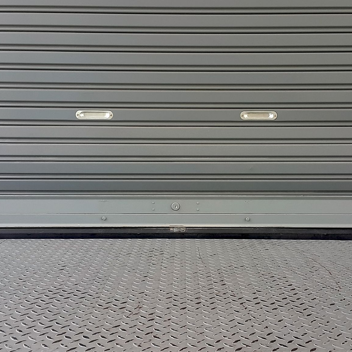 Porte enroulable en métal gris, fermée, avec deux lumières rectangulaires au-dessus d'un sol gris texturé.