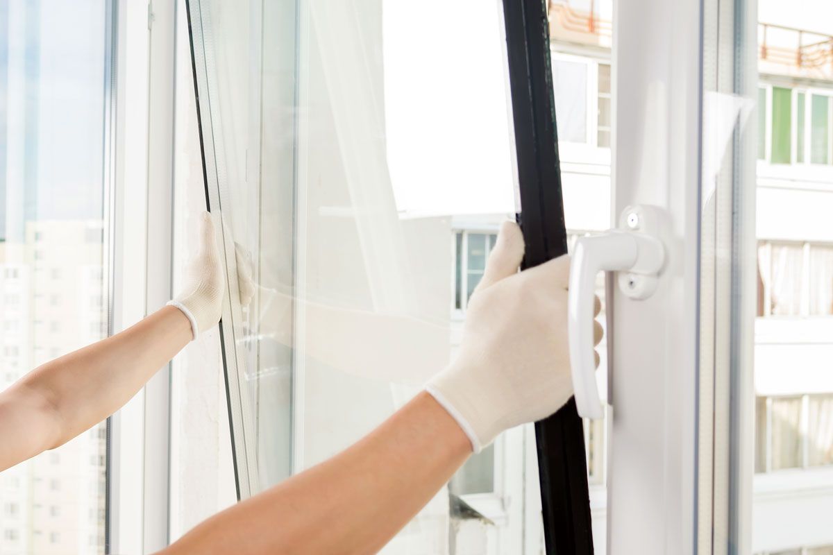 Une personne portant des gants installe un joint noir sur un cadre de fenêtre blanc à l'intérieur.