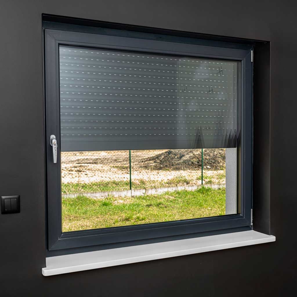 Fenêtre avec cadre gris foncé et volet roulant, couvrant partiellement la vue sur la zone herbeuse et le chantier.