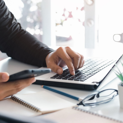 Personne tapant sur un ordinateur portable avec un téléphone dans une main ; bureau avec cahier, crayon et lunettes.