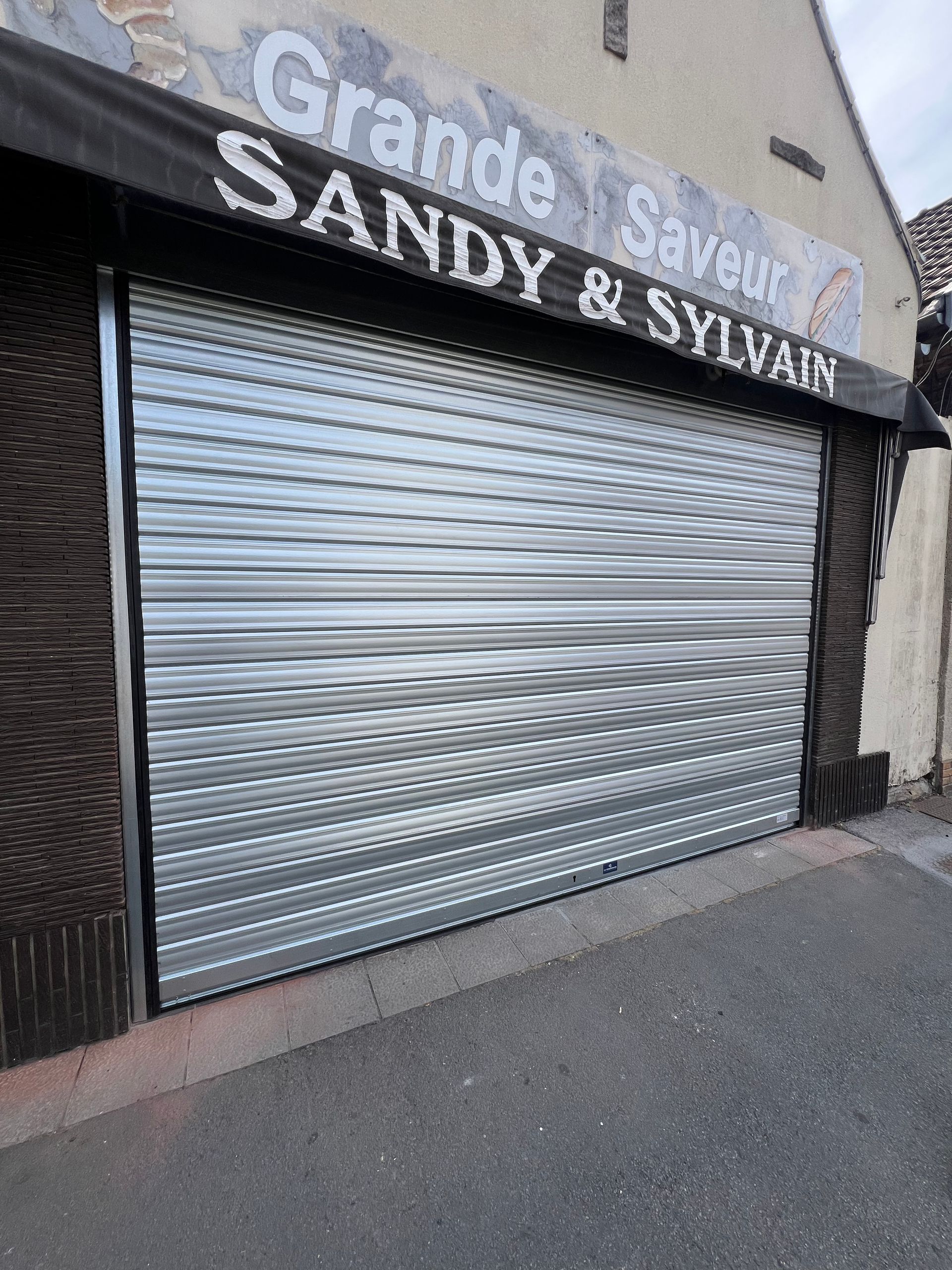 Façade de magasin fermée avec rideau métallique argenté ; enseigne « Grande Saveur SANDY & SYLVAIN » au-dessus.