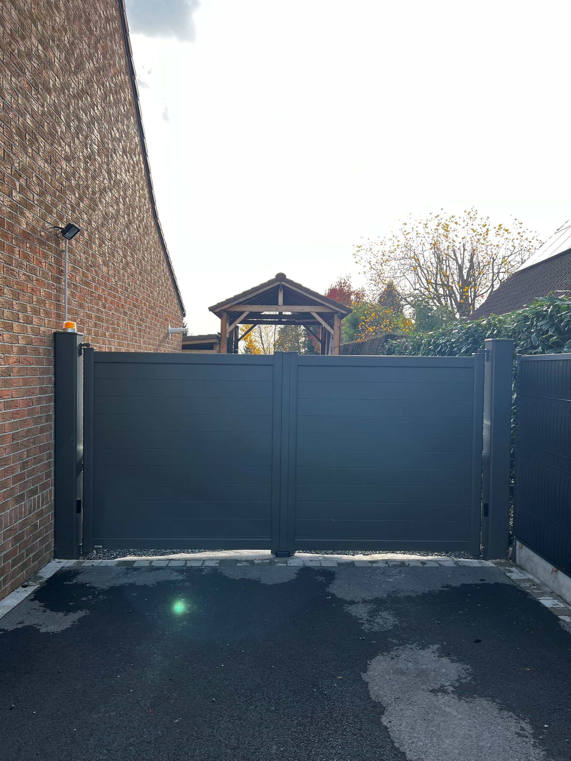 Un portail métallique gris foncé, fermé, se dresse devant un bâtiment en briques. À l'arrière-plan, on aperçoit un kiosque et des arbres.