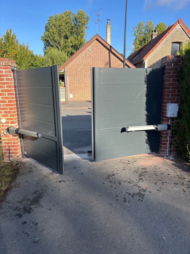 Portail métallique gris, ouvert, avec des piliers en briques, devant une maison de campagne.