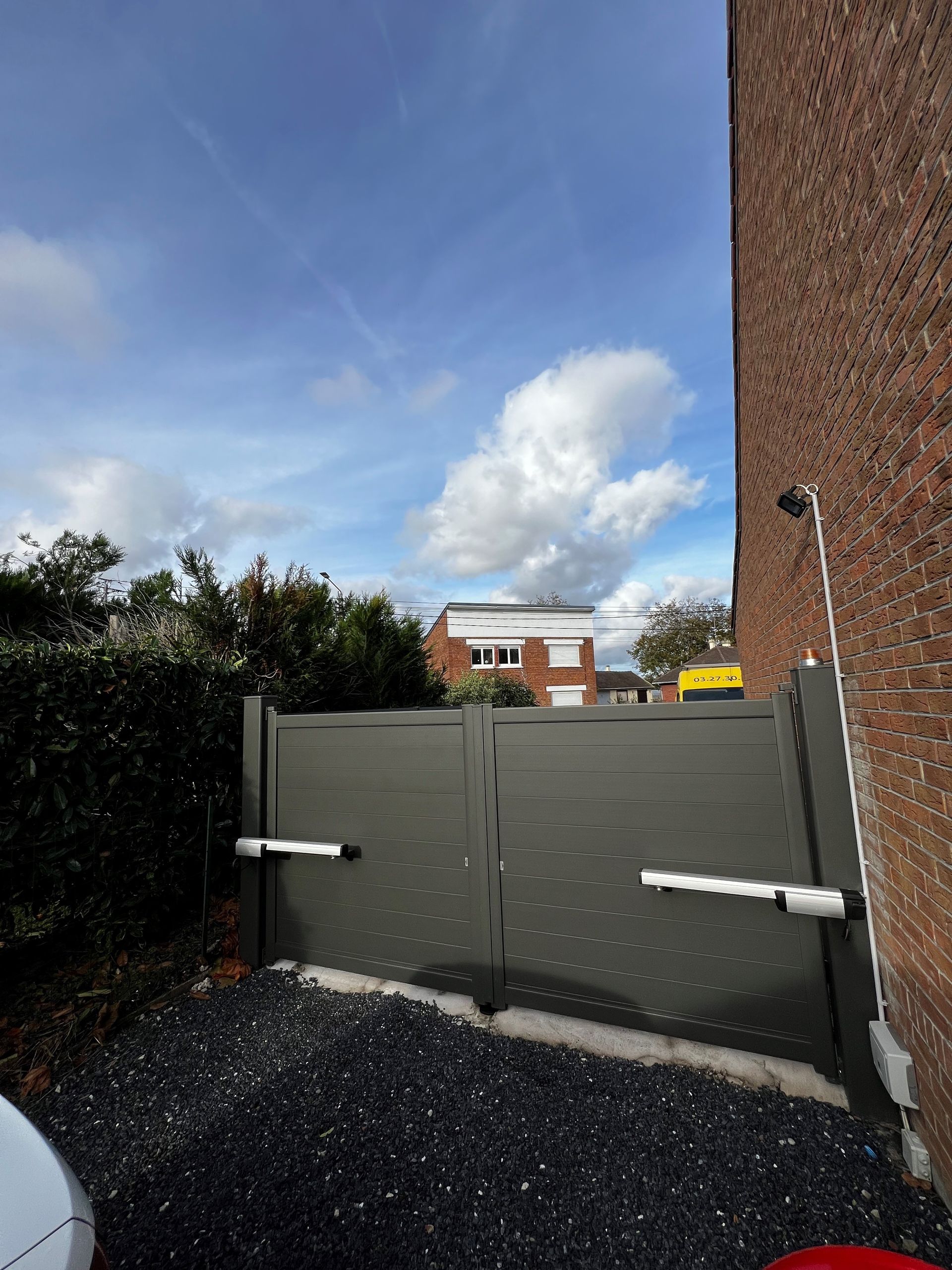 Portail gris foncé, gravier noir, ciel bleu avec des nuages blancs, bâtiment en briques à droite, buissons verts.