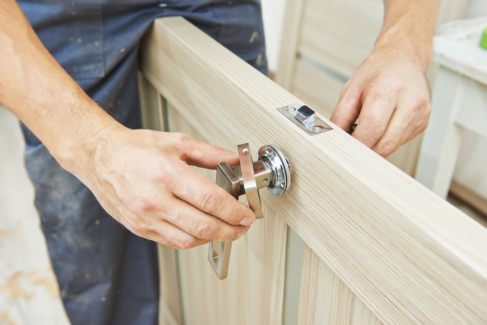 Une personne en tenue de travail installe une poignée de porte sur une porte en bois clair.