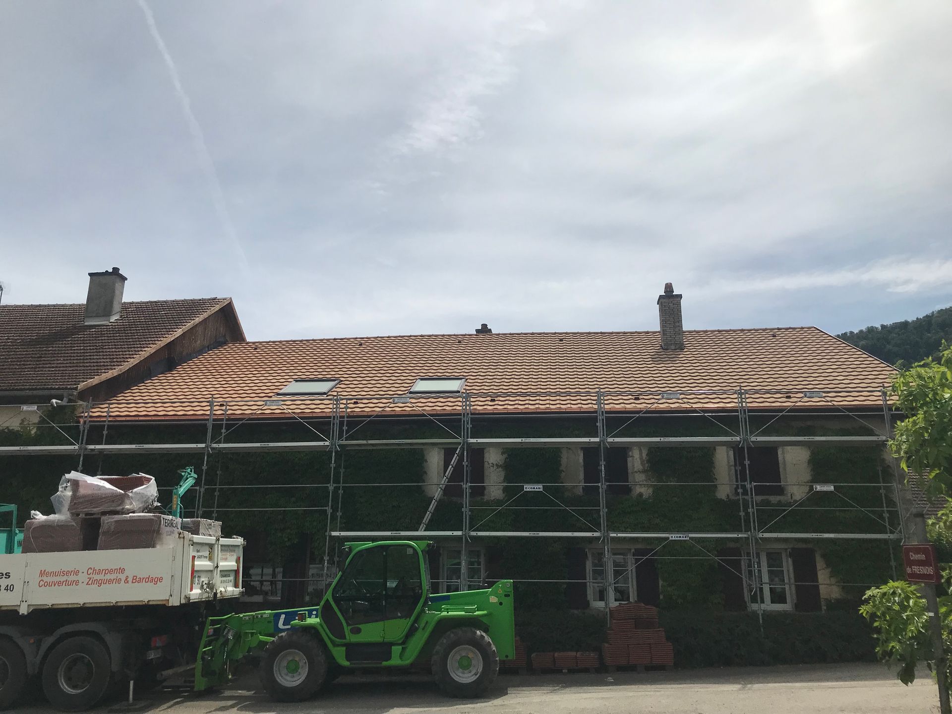 Des échafaudages entourent une maison dont le toit de tuiles affaissé est en cours de réparation, un chariot télescopique vert étant garé devant.
