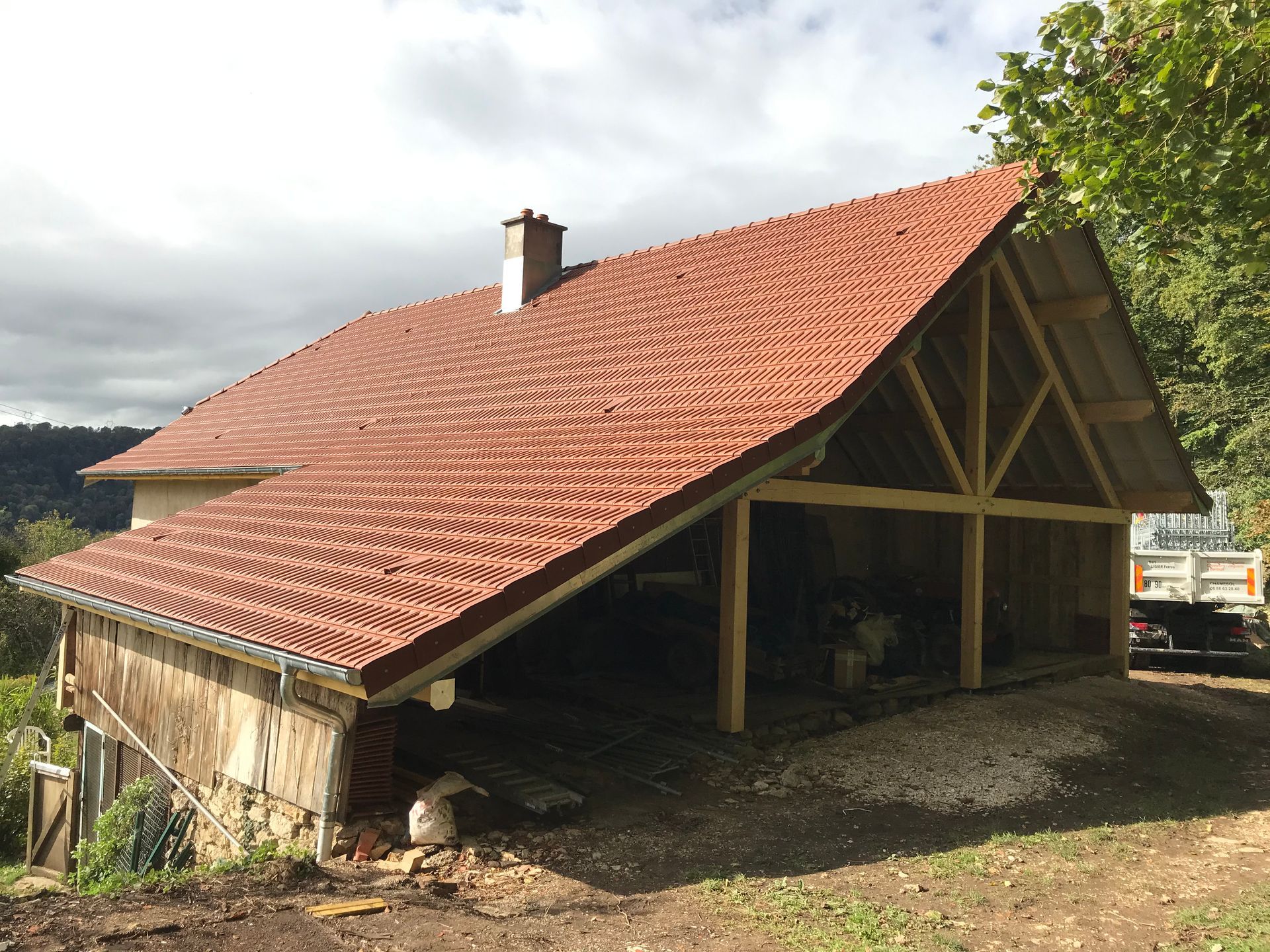 Une maison couleur pêche avec un toit de tuiles brun foncé, un puits de lumière et une cheminée, partiellement dissimulée par une haie verte.