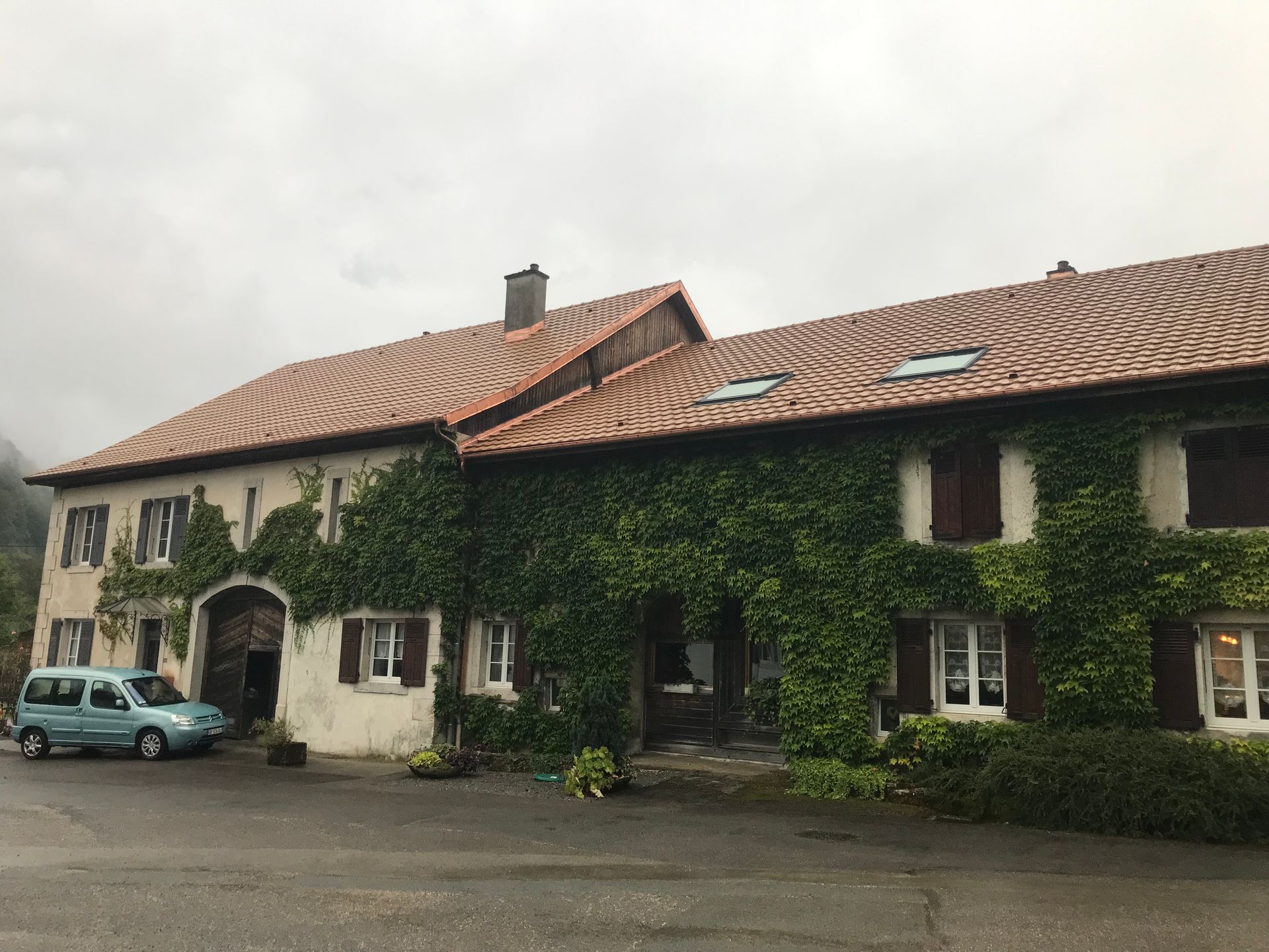 Une grande ferme en pierre à deux étages, au toit de tuiles recouvert de lierre grimpant, avec une voiture bleu clair garée devant.