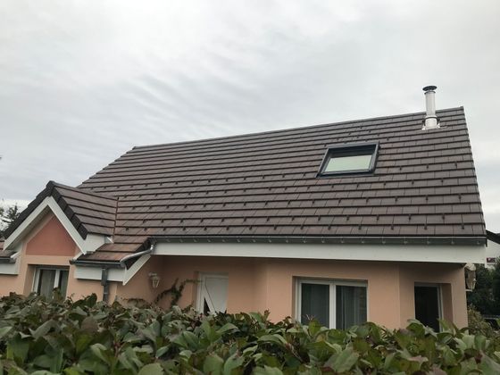 Une maison couleur pêche avec un toit de tuiles brun foncé, un puits de lumière et une cheminée, partiellement dissimulée par une haie verte.