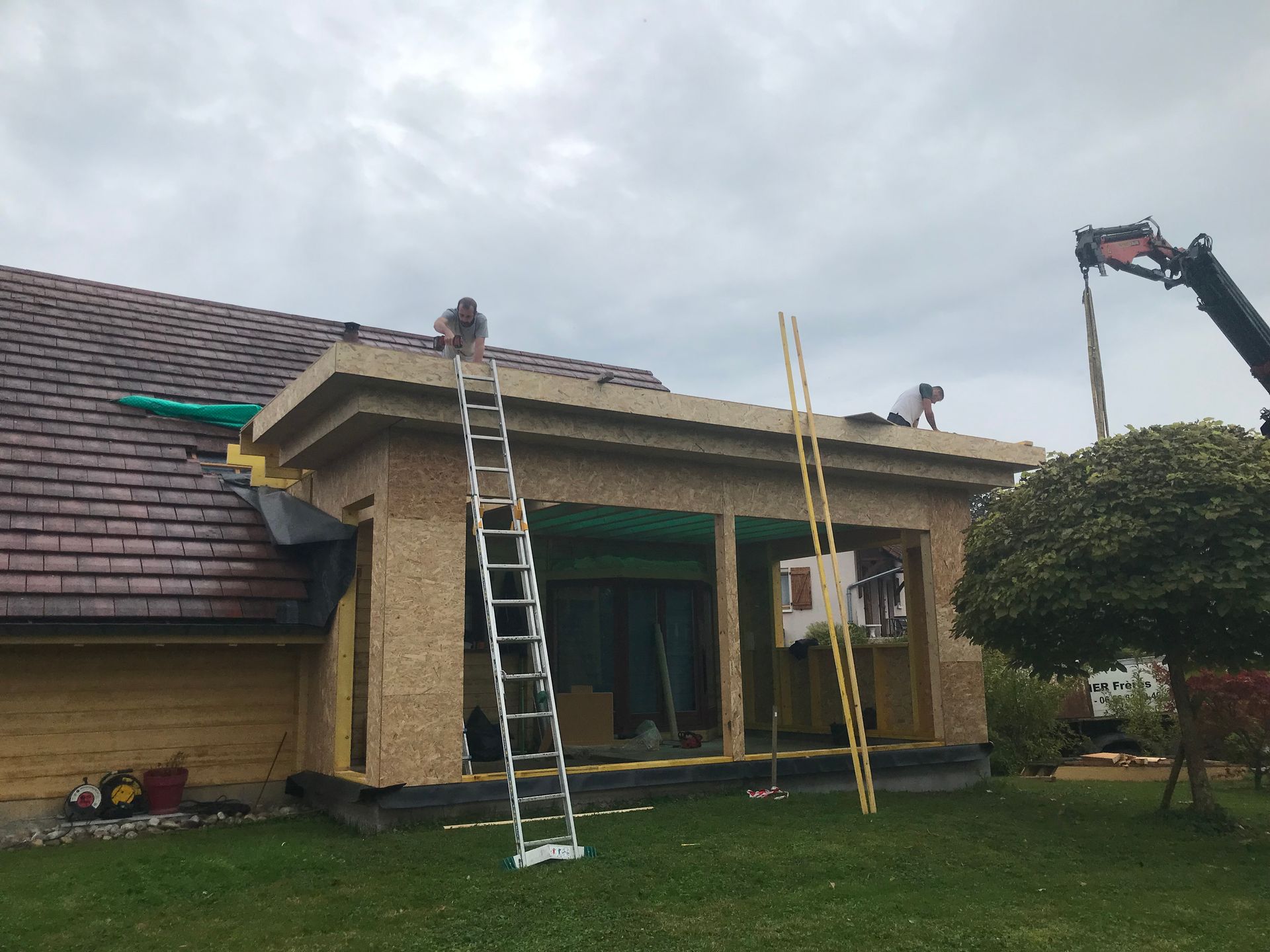 Deux ouvriers, juchés sur des échelles, rénovent une extension de maison en bois sous un ciel nuageux, avec une grue visible en arrière-plan.