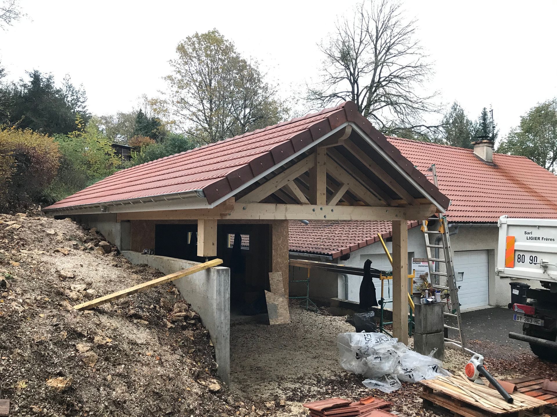 Un abri de voiture en bois inachevé, au toit de tuiles rouges, adossé à une maison au milieu d'un chantier.