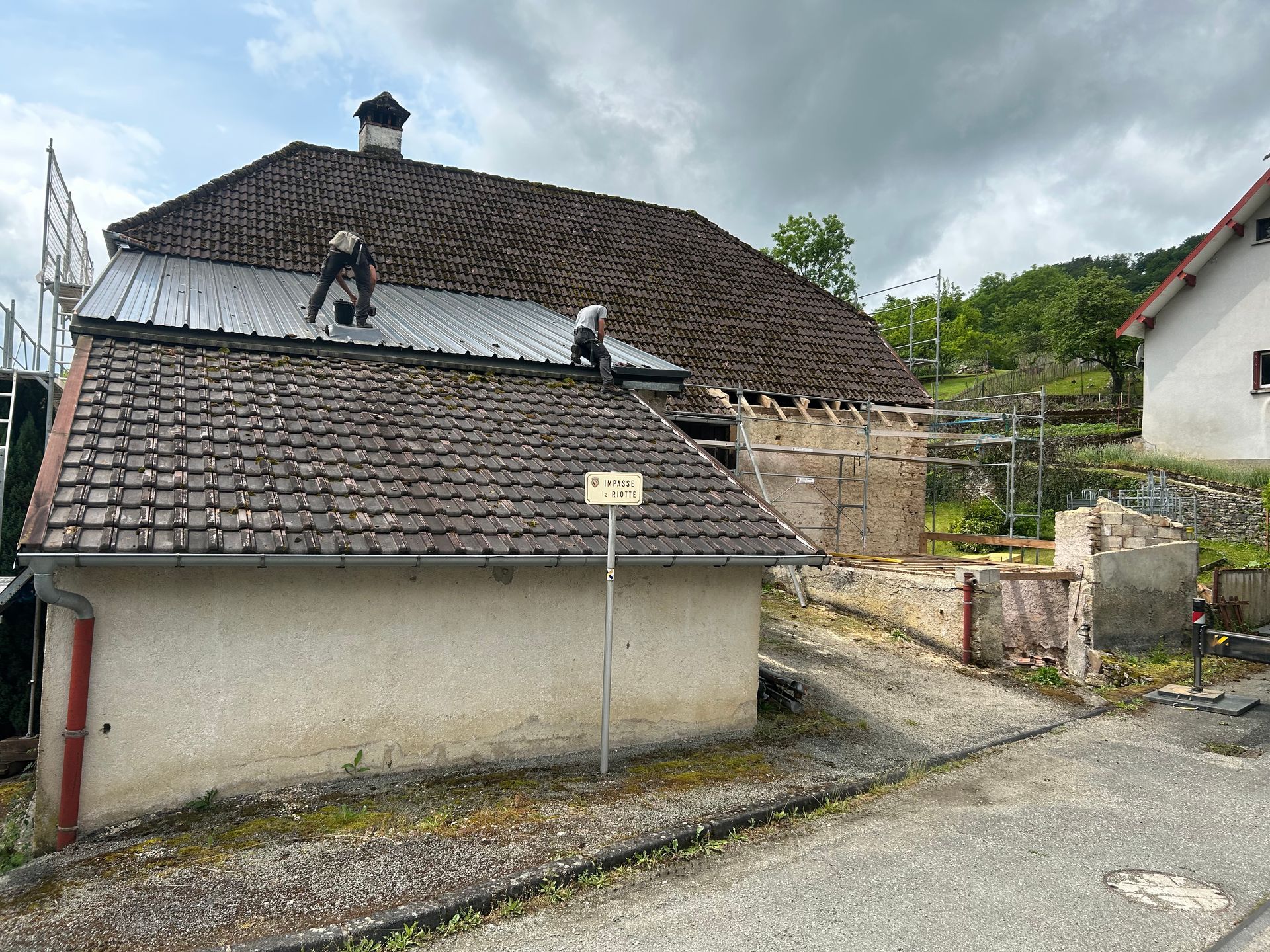 Deux ouvriers installent une nouvelle section de toiture métallique sur un vieux bâtiment partiellement en ruine, recouvert d'une toiture en tuiles traditionnelles.