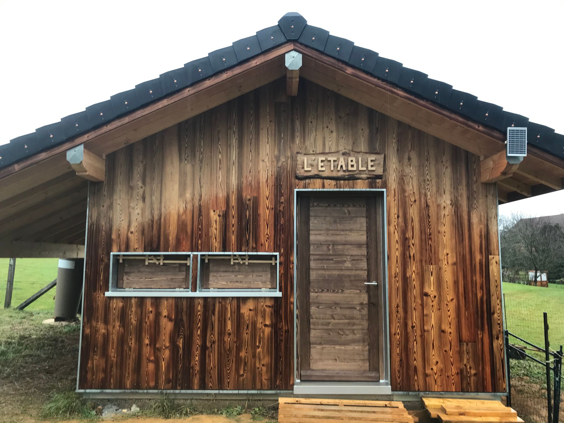 Un bâtiment rustique en bois portant l'inscription L'ETABLE, avec une porte centrale et deux fenêtres latérales, situé dans un espace extérieur gazonné.
