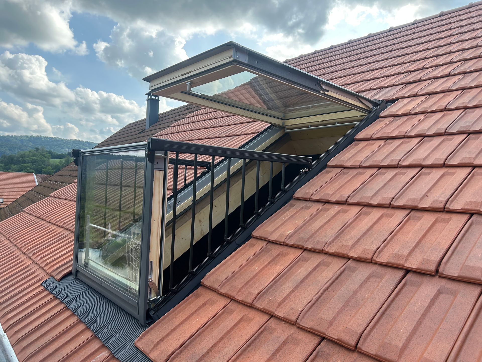 Une fenêtre de toit ouverte en configuration balcon, avec une façade vitrée et une rambarde noire, sur un toit de tuiles rouges.